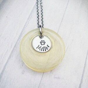 May include: A silver necklace with a circular pendant and a smaller, round charm. The smaller charm has a paw print and the name "Miller" in black. The larger pendant is a light yellow colour.