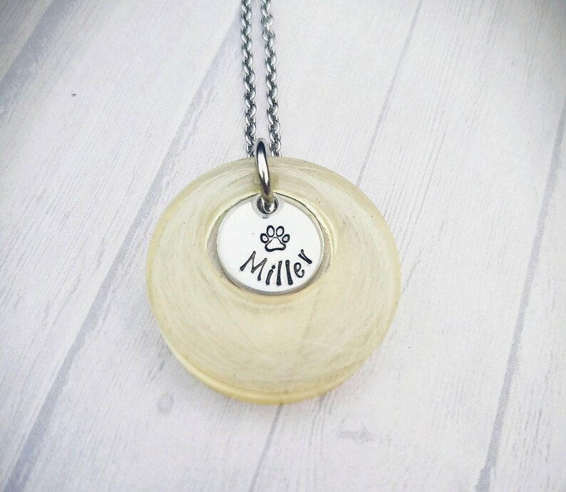 May include: A silver necklace with a circular pendant and a smaller, round charm. The smaller charm has a paw print and the name "Miller" in black. The larger pendant is a light yellow colour.