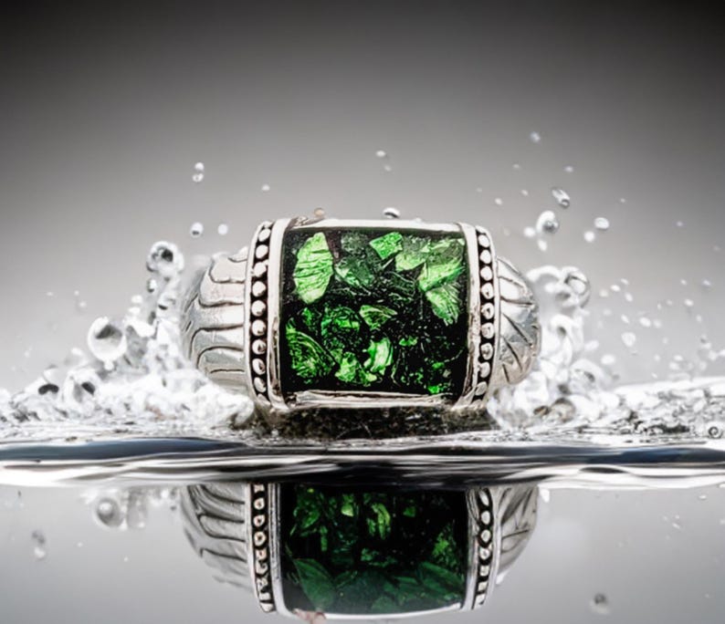 May include: A silver ring with a green gemstone set in a silver bezel. The ring is surrounded by water droplets.