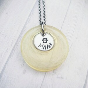May include: A silver necklace with a circular, pale yellow pendant. A smaller, silver disc is attached, with a paw print and the name "Miller". The necklace chain is also silver.