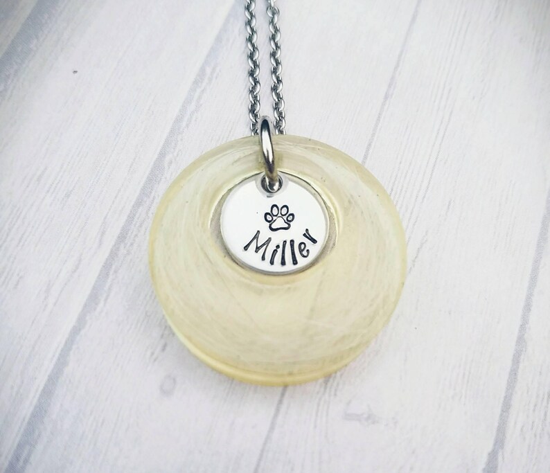 May include: A silver necklace with a circular, pale yellow pendant. A smaller, silver disc is attached, with a paw print and the name "Miller". The necklace chain is also silver.