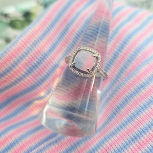 May include: A silver ring with a square, opalescent stone, surrounded by a halo of small, clear stones. The ring is displayed on a clear, conical stand against a backdrop of pink and blue striped fabric.