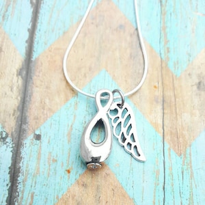 May include: A silver-colored pendant necklace featuring a teardrop-shaped urn and a cut-out angel wing charm. The urn has a small, decorative crystal at the bottom. The necklace is displayed on a blue and brown chevron patterned surface.