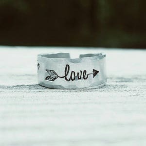 May include: A silver-colored ring with the word "love" and an arrow design. The ring has a hammered texture and is set against a blurred background.