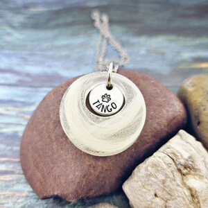 May include: A silver pendant necklace with a circular design. The pendant has a smaller disc with the word "TANGO" and a paw print. The necklace is displayed on a brown rock.