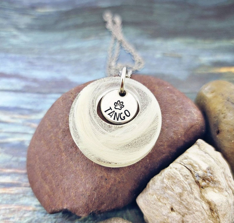 May include: A silver pendant necklace with a circular design. The pendant has a smaller disc with the word "TANGO" and a paw print. The necklace is displayed on a brown rock.