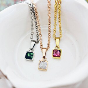May include: Three delicate necklaces with square pendants featuring different colored gemstones. The pendants are silver, gold, and rose gold. The gemstones are green, pink, and clear.