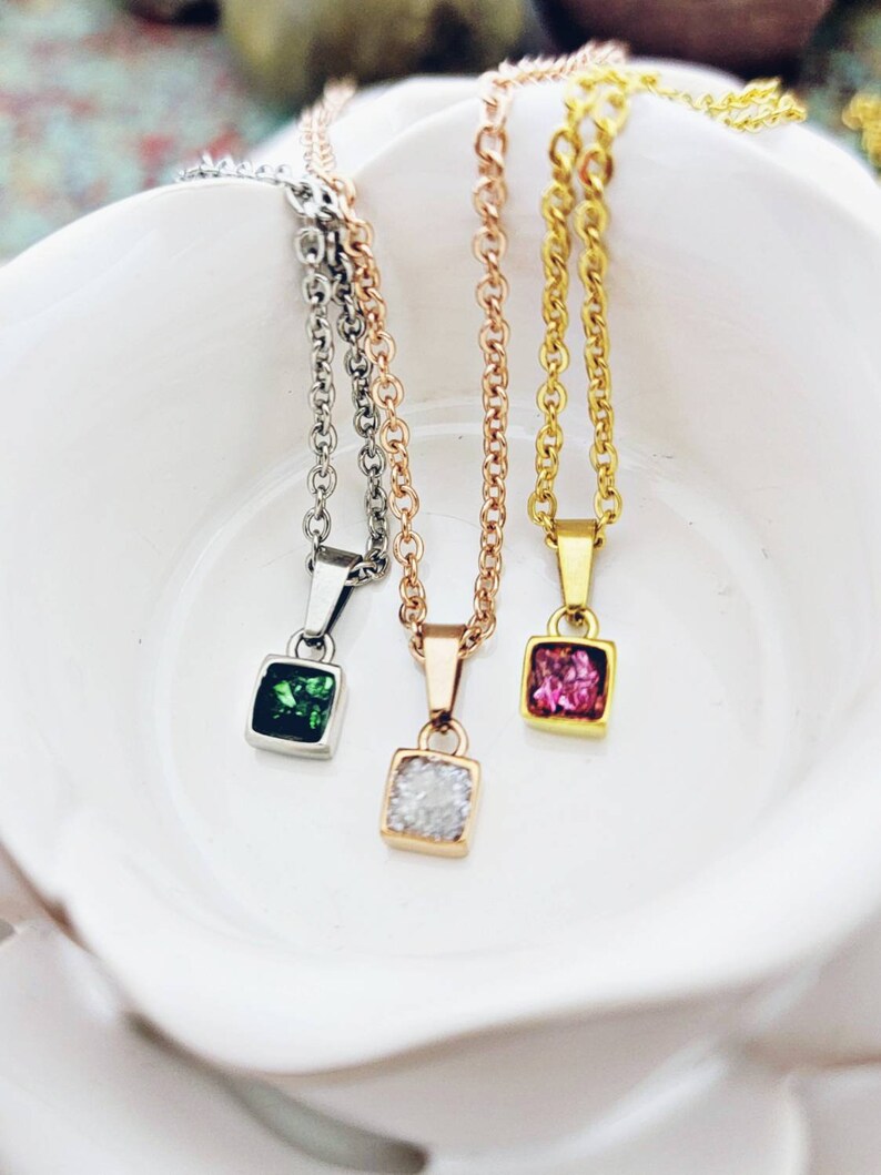 May include: Three delicate necklaces with square pendants featuring different colored gemstones. The pendants are silver, gold, and rose gold. The gemstones are green, pink, and clear.