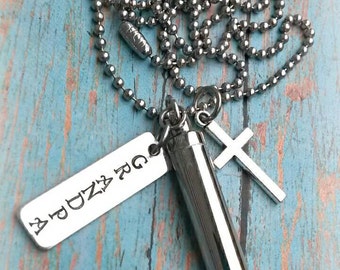 May include: Silver necklace with a small silver cross charm, a silver tag engraved with the text "BUDDY RG", and a silver cylinder charm. The necklace is on a blue wooden background.