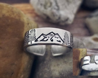 Puede incluir: Un anillo de plata con un diseño grabado de montañas y árboles. El anillo se muestra sobre una superficie de roca marrón.
