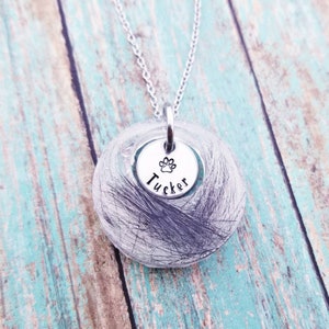 May include: A silver necklace featuring a clear, circular pendant filled with dark grey strands. A smaller silver disc with a paw print and the name "Tucker" is attached. The necklace hangs on a silver chain.