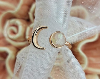 May include: Gold-toned ring featuring a crescent moon and a round, iridescent stone. The ring's design includes an open band, with the moon and stone positioned on either side. The ring is set against a backdrop of white fabric and soft, blurred colors.