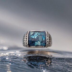 May include: A silver ring with a blue and green gemstone set in a square shape. The ring is surrounded by a silver band with a textured design.
