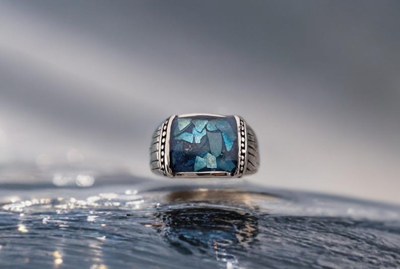 May include: A silver ring with a blue and green gemstone set in a square shape. The ring is surrounded by a silver band with a textured design.