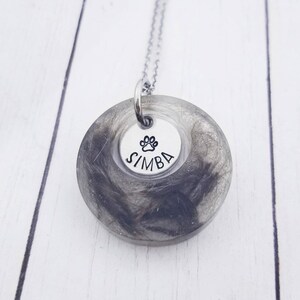 May include: A pendant necklace with a silver chain. The pendant has a clear resin circle filled with grey and black hair. A smaller silver disc in the centre has a paw print and the name "SIMBA".