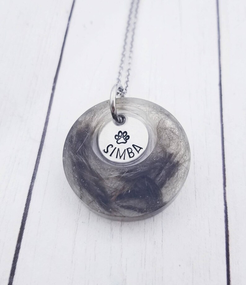 May include: A pendant necklace with a silver chain. The pendant has a clear resin circle filled with grey and black hair. A smaller silver disc in the centre has a paw print and the name "SIMBA".
