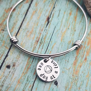 May include: Silver bangle bracelet with a round charm that says "one day at a time" and features a triangle symbol.