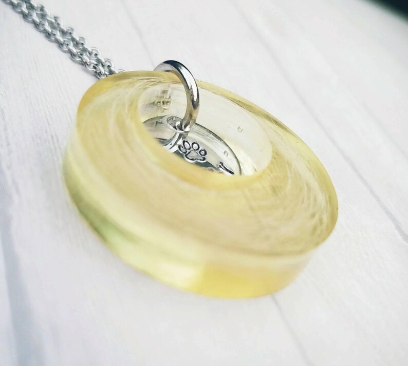 May include: A pendant necklace with a clear, oval resin, displaying a yellow and white swirl pattern. Inside the resin is a small, silver charm with a paw print. The necklace has a silver chain.