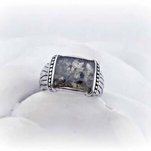 May include: A silver ring with a rectangular gray stone set in the center. The stone has a speckled pattern and is surrounded by a beaded border.