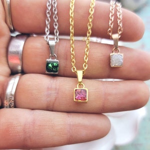 May include: Three delicate necklaces with square-shaped pendants. The pendants are made of clear, white, green, and pink crystals. The necklaces are on a gold, silver, and rose gold chain.