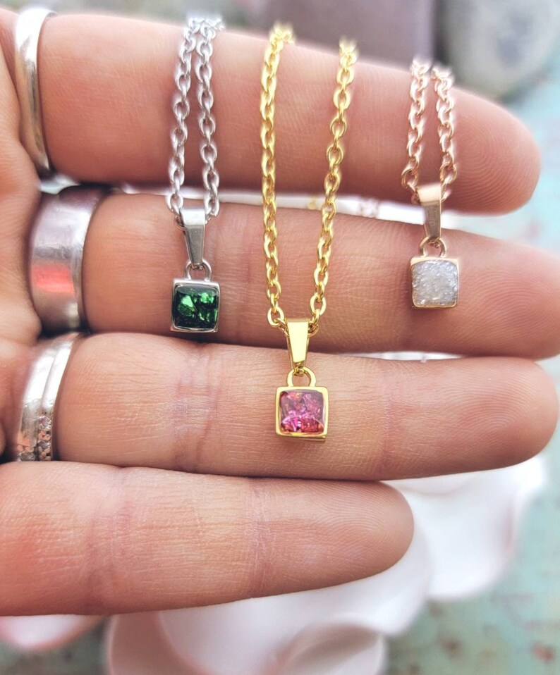 May include: Three delicate necklaces with square-shaped pendants. The pendants are made of clear, white, green, and pink crystals. The necklaces are on a gold, silver, and rose gold chain.