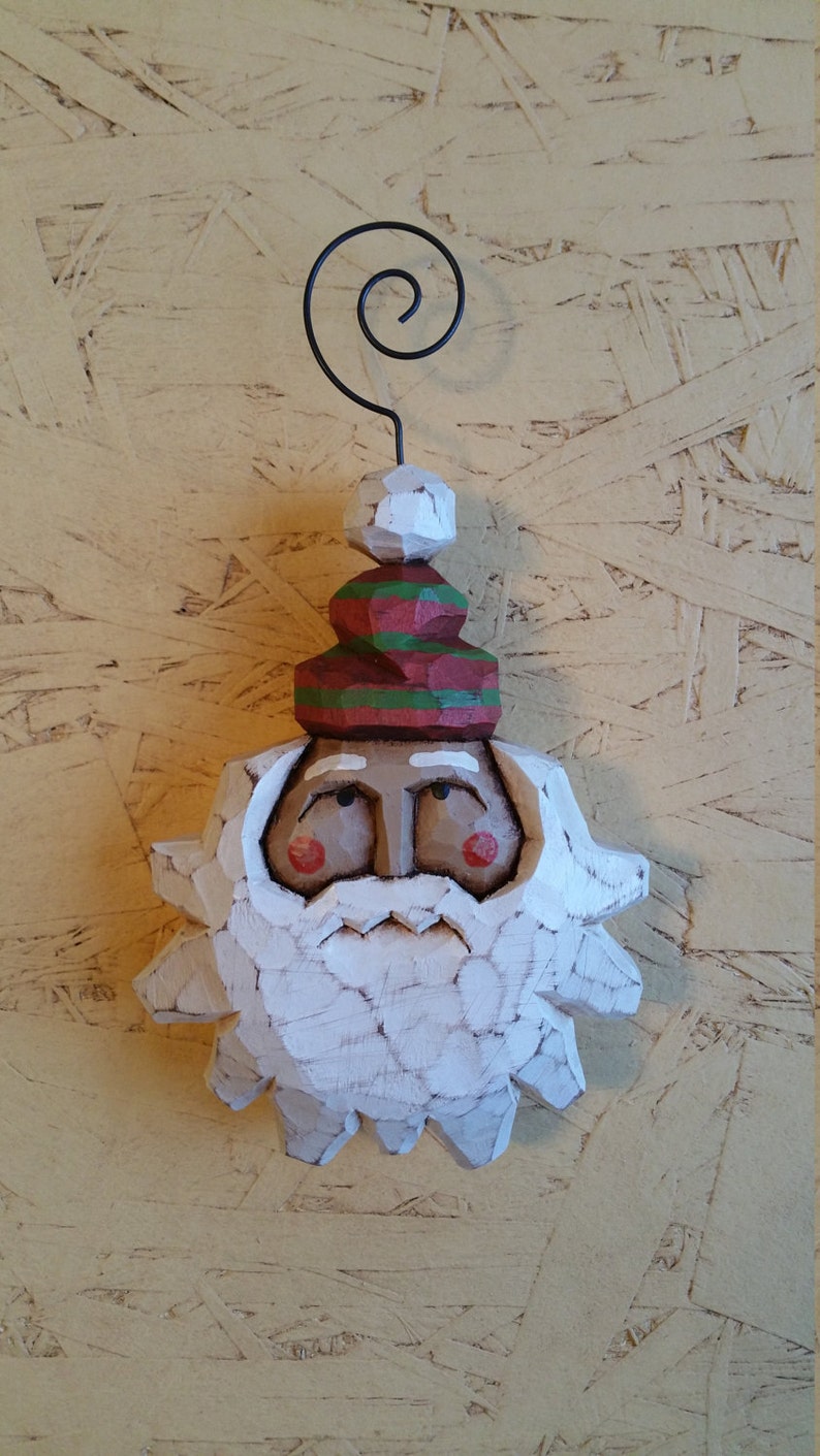 May include: A carved wooden Santa Claus ornament with a white beard and a red and green hat. The ornament has a wire loop for hanging. The background is a light brown wood.
