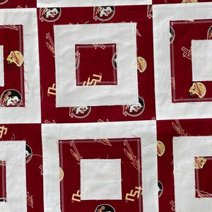 May include: A white and red fabric quilt with a repeating pattern of a Seminole State University logo. The quilt is made up of squares with a white background and a red border. The squares are arranged in a grid pattern.