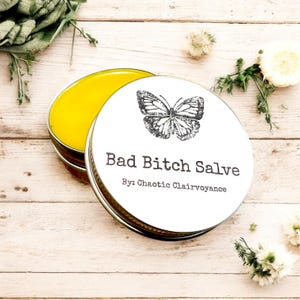 May include: A round metal tin with a white lid featuring a butterfly illustration and the text "Bad Bitch Salve" with "By: Chaotic Clairvoyance" below. The tin is open, revealing a yellow salve. The background is a light wood surface.