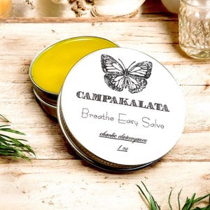 May include: A round, open metal tin filled with yellow salve, next to a lid with a butterfly illustration and the text "CAMPAPAKALATA Breathe Easy Salve". The lid also includes the text "chaotic clairvoyance" and "1 oz".