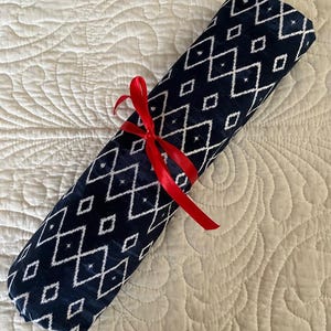May include: Rolled navy blue fabric with a white diamond pattern, tied with a red ribbon. The fabric is rolled up and appears to be a scarf or a similar textile item. The background is a cream-colored, quilted surface.