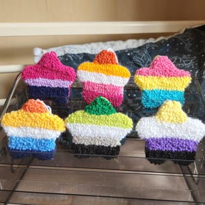May include: Six star-shaped punch needle ornaments arranged on a clear tiered display stand. Each star features a unique horizontal colour pattern, including combinations of pink, purple, dark blue, orange, yellow, white, green, grey, and black yarn.