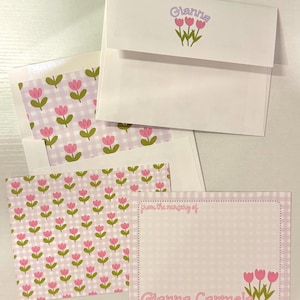 May include: Stationery set featuring white envelopes with the name "Gianna" and pink tulip illustrations. The set includes notecards with a pink gingham pattern and matching tulip designs. The phrase "from the nursery of" is also printed on the notecards.