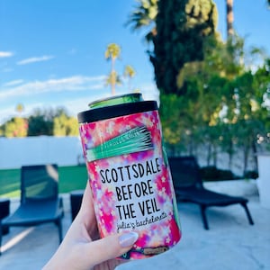 Puede incluir: Un enfriador de latas cilíndrico con un vibrante patrón de estrellas rosas, moradas y blancas. Se muestra el texto "SCOTTSDALE BEFORE THE VEIL julia's bachelorette". Una lata verde está dentro. El fondo incluye palmeras y un cielo azul.