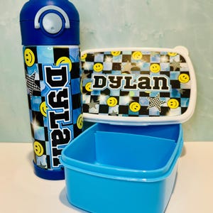 Puede incluir: Un juego de fiambrera azul y negro con un patrón a cuadros y caras sonrientes. La fiambrera tiene el nombre "Dylan" impreso en ella. El juego incluye una botella de agua con una tapa azul y un diseño a juego.