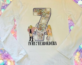 Puede incluir: Una camiseta blanca de manga larga con un gráfico grande del número siete relleno de confeti multicolor. El diseño incluye dos figuras estilizadas y el texto negro IN MY 7 YEAR OLD ERA impreso debajo del gráfico del número central.