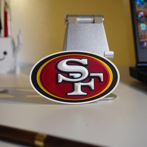 Può includere: Un emblema ovale con le lettere "SF" bianche su sfondo rosso, bordato di nero e oro. Questo è probabilmente un logo di una squadra sportiva, forse di San Francisco.