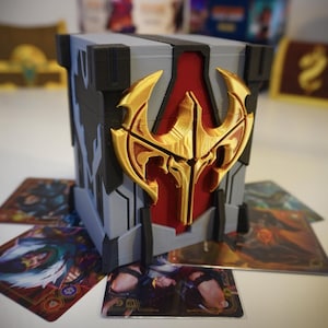May include: A gray and black cube-shaped box with a gold emblem and red accents, surrounded by trading cards. The emblem is a stylized design. The cards feature colorful illustrations.