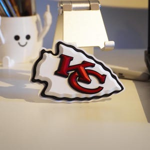 May include: A white arrowhead-shaped emblem with a black outline and the red letters "KC" in the centre. The emblem is on a white surface, with a laptop and a pen holder in the background.