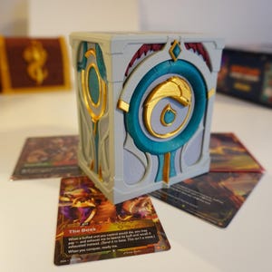 May include: A gray, teal, and gold decorative box with a stylized design, resting on trading cards. The box features a central teal circle with a gold emblem. Other boxes and a blue box are in the background.