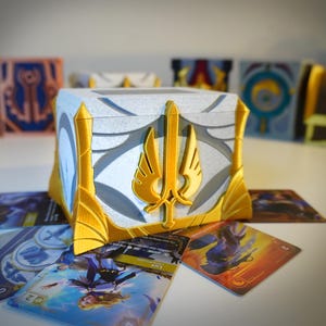 May include: A white and gold decorative box with a sword and wing design, surrounded by trading cards. The box has a textured surface and is accented with gold trim. Other decorative boxes are in the background.