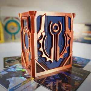 Puede incluir: Una caja geométrica de color cobre con intrincados diseños recortados, sobre un fondo azul. La caja está rodeada de cartas coleccionables. Otras cajas son visibles en el fondo.