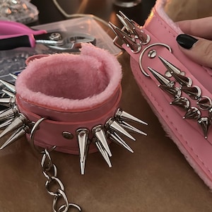Op de afbeelding: Roze leren boeien met zilveren spikes en een zachtroze binnenkant. Inclusief een bijpassende polsband en een ketting. De polsband en kraag zijn versierd met meerdere rijen zilveren spikes.