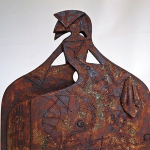 Puede incluir: Una escultura de cerámica de una figura estilizada con un esmalte de color óxido. La figura está de pie con los brazos levantados y tiene un diseño geométrico en su cuerpo.