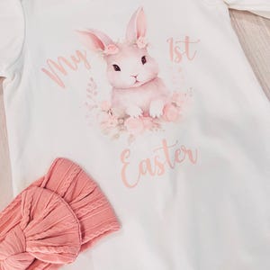 K&ouml;nnte beinhalten: Wei&szlig;er Langarm-Strampler mit einem rosa Hasen, der eine Blumenkrone tr&auml;gt und den Text "My 1st Easter". Ein rosa Stirnband ist ebenfalls enthalten.