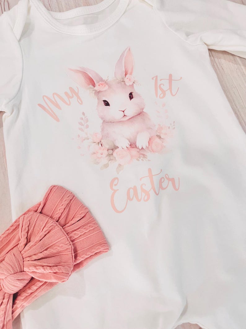 K&ouml;nnte beinhalten: Wei&szlig;er Langarm-Strampler mit einem rosa Hasen, der eine Blumenkrone tr&auml;gt und den Text "My 1st Easter". Ein rosa Stirnband ist ebenfalls enthalten.