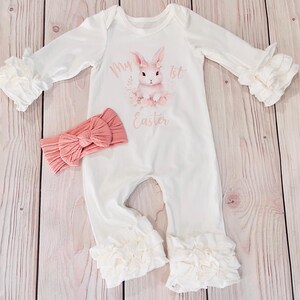 K&ouml;nnte beinhalten: Wei&szlig;er Baby-Strampler mit einem rosa Hasen und dem Text "My 1st Easter". Der Strampler hat R&uuml;schen an den &Auml;rmeln und Beinen. Ein rosa Stirnband ist im Lieferumfang enthalten.