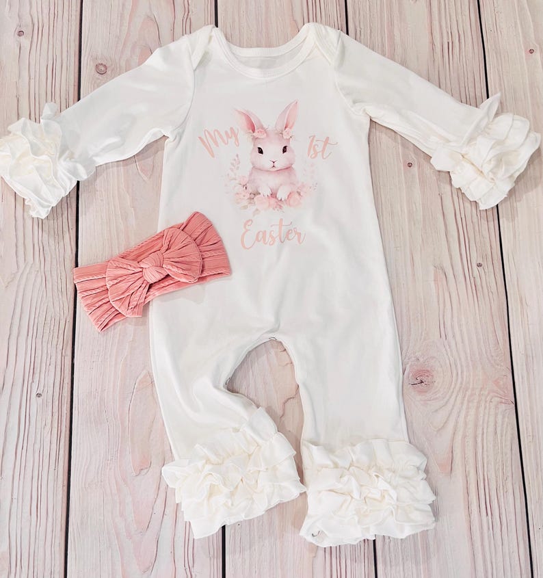 K&ouml;nnte beinhalten: Wei&szlig;er Baby-Strampler mit einem rosa Hasen und dem Text "My 1st Easter". Der Strampler hat R&uuml;schen an den &Auml;rmeln und Beinen. Ein rosa Stirnband ist im Lieferumfang enthalten.