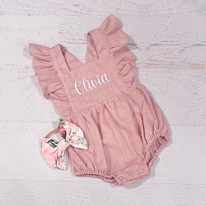 May include: A dusty rose baby romper with ruffled straps and the name "Olivia" in white script. A matching floral bow headband is next to the romper. The romper has a bubble silhouette and snap closures.