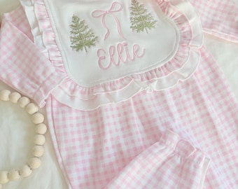 Könnte beinhalten: Ein langärmeliger Baby-Strampler mit hellrosa-weißem Vichy-Muster und einem weißen, gerüschten Lätzchenkragen. Der Kragen ist mit zwei grünen Tannenbäumen, einer rosa Schleife und dem Namen Ellie in rosa Schreibschrift bestickt. Eine große rosa Haarschleife liegt bei.