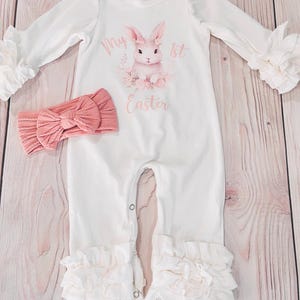 K&ouml;nnte beinhalten: Wei&szlig;er Baby-Strampler mit einem rosa Hasen-Motiv und dem Text "My 1st Easter". Der Strampler hat R&uuml;schen an den &Auml;rmeln und Beinen. Ein rosa Stirnband ist im Lieferumfang enthalten.
